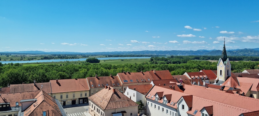 An aerial view of Brezice with its traditional red-tiled roofs and a St. Lawrence's Parish Church steeple, overlooking the green landscape with the Sava river in the distance