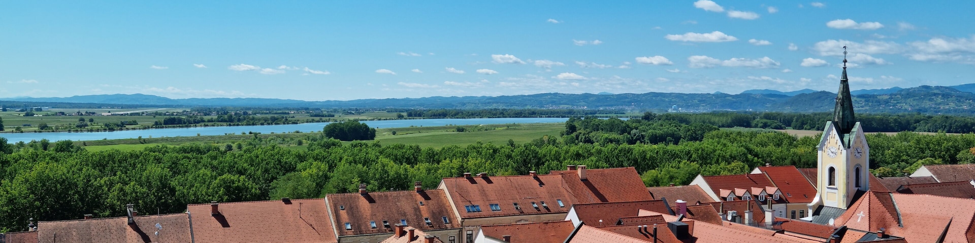 An aerial view of Brezice with its traditional red-tiled roofs and a St. Lawrence's Parish Church steeple, overlooking the green landscape with the Sava river in the distance