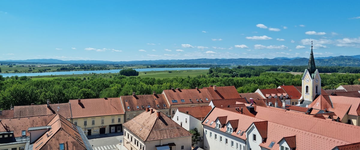 A town with red-roofed buildings, a church with a clock tower, and a river in the background.