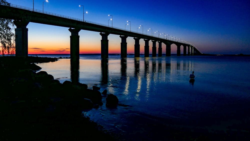 Eine lange Brücke in der Dämmerung mit Lichtern, die sich über ruhiges Wasser spannt.