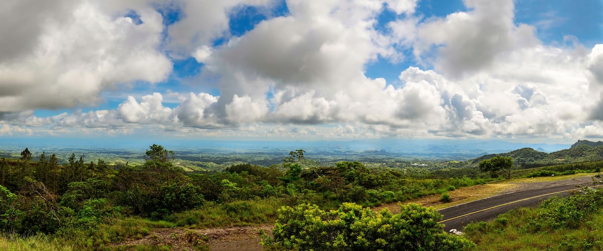 A winding road through lush green hills with a distant view of a valley and mountains under a partly cloudy sky.
