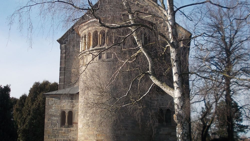 A historic stone church with a bell tower, surrounded by a cemetery with headstones and a large tree in the foreground.