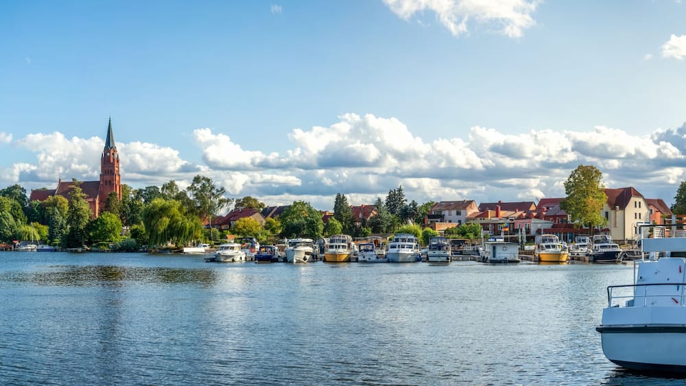 A calm river with boats docked, a church with a tall spire, and residential buildings along the riverbank.