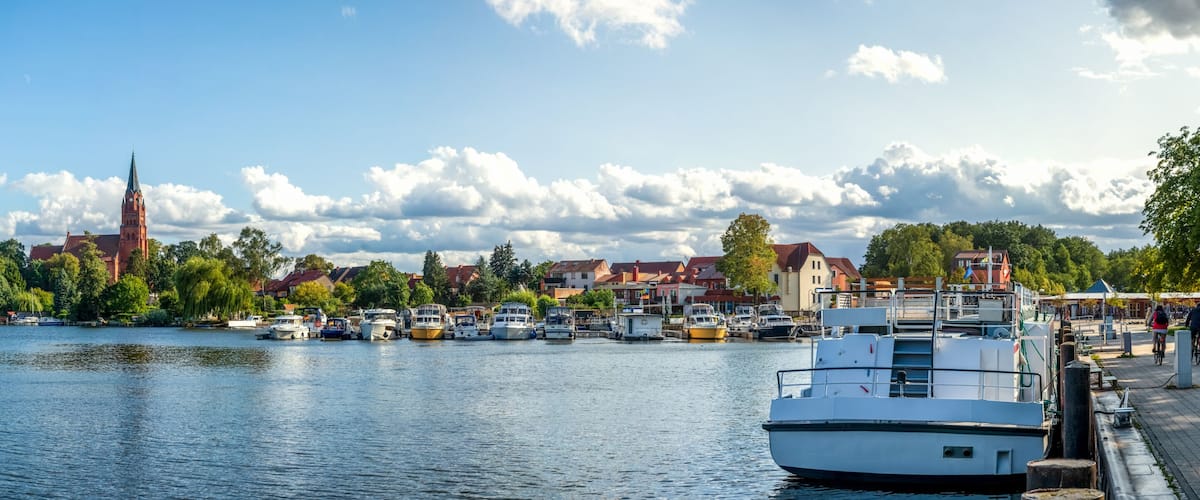A calm river with boats docked, a church with a tall spire, and residential buildings along the riverbank.