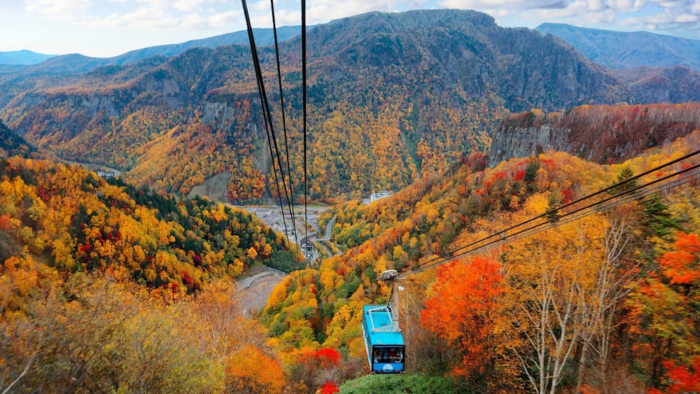 Eine Seilbahn bewegt sich durch eine gebirgige Landschaft mit Herbstlaub.