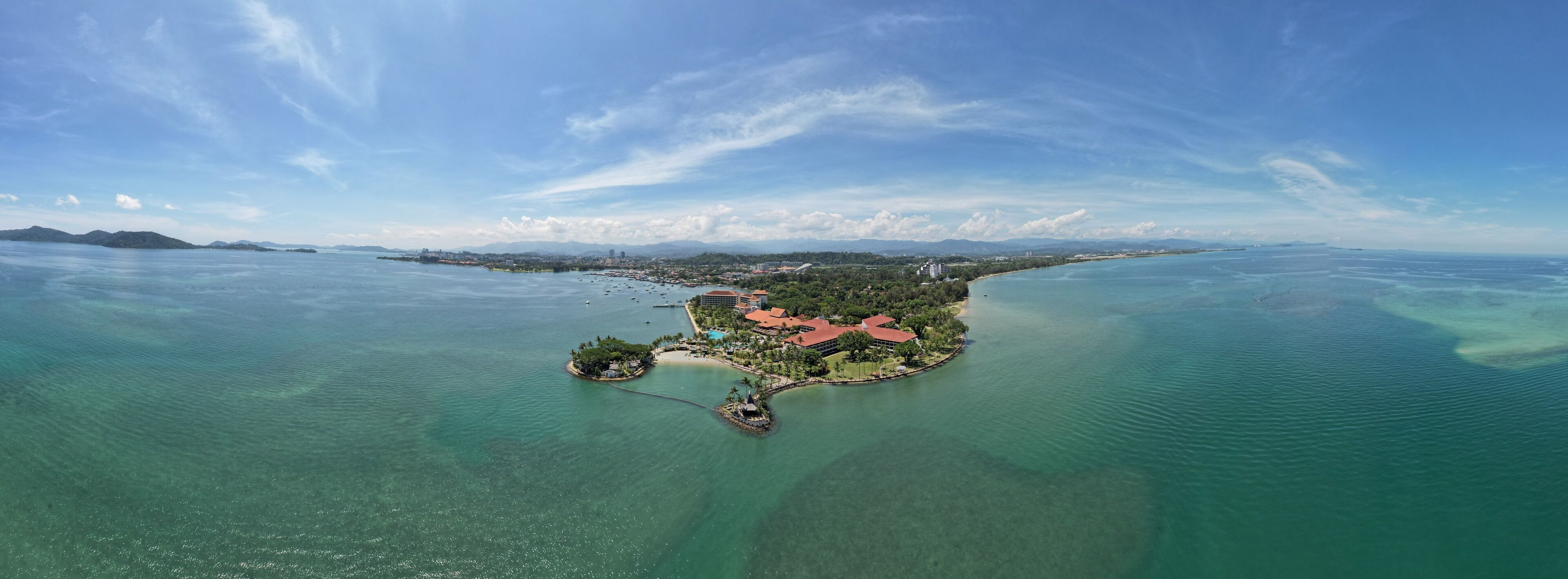 An aerial view of a tropical island with clear turquoise waters, a resort area with buildings, and a distant coastline.