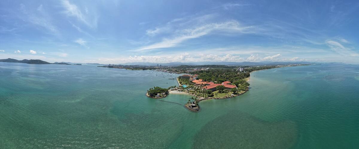 An aerial view of a tropical island with clear turquoise waters, a resort area with buildings, and a distant coastline.
