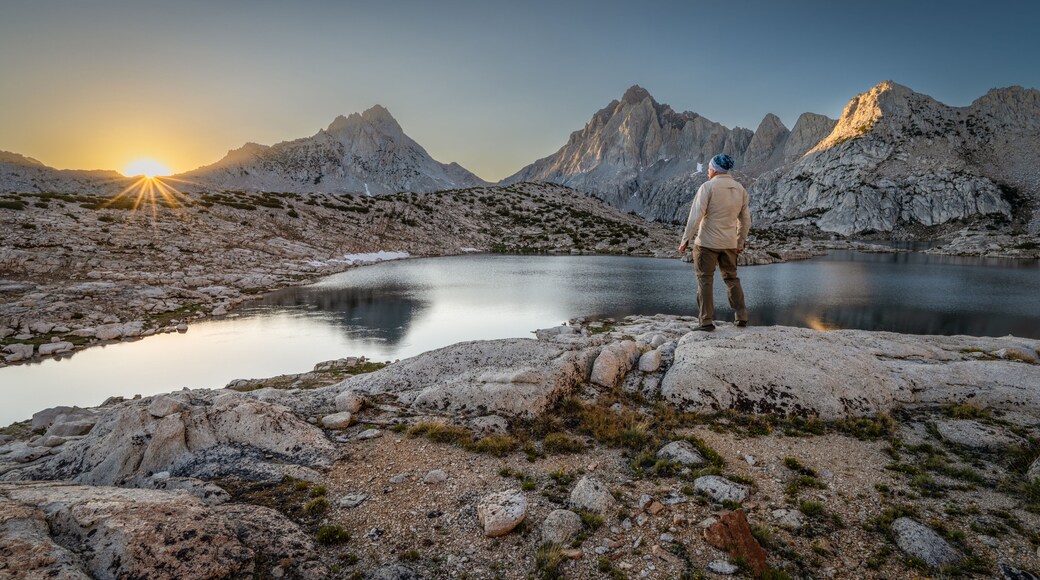 Rear view of a man standing on a rock in front of Grinnell lake at dawn looking at the Sierra Nevada Mountains, Sierra National Forest, California, USA