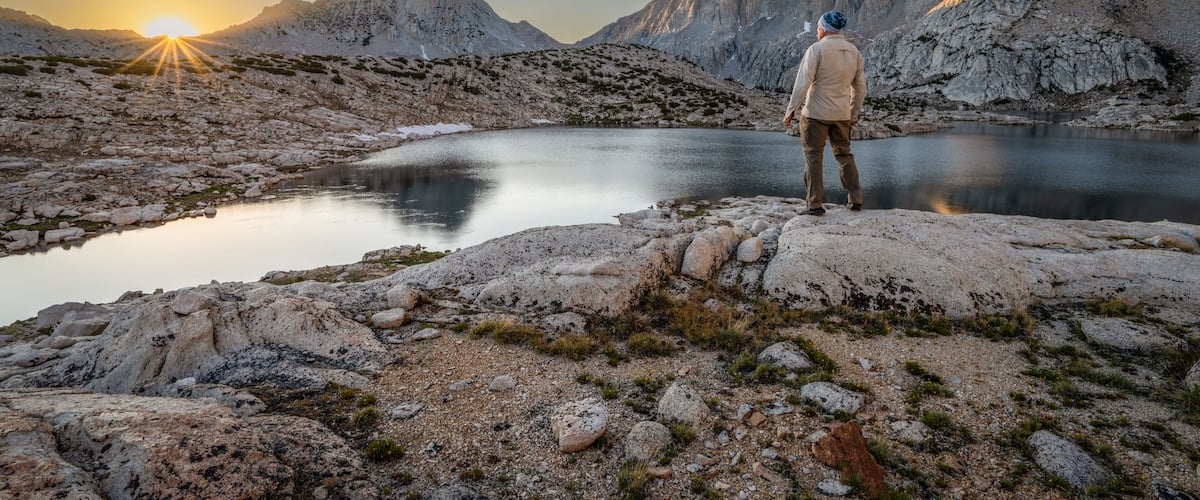 Rear view of a man standing on a rock in front of Grinnell lake at dawn looking at the Sierra Nevada Mountains, Sierra National Forest, California, USA