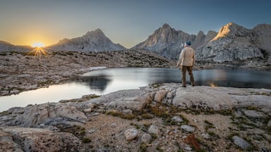 Rear view of a man standing on a rock in front of Grinnell lake at dawn looking at the Sierra Nevada Mountains, Sierra National Forest, California, USA