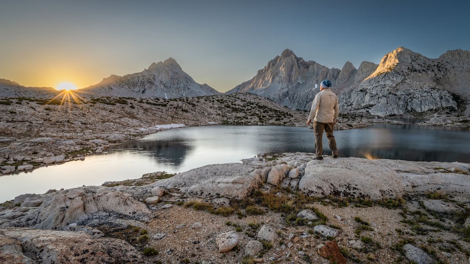 Rear view of a man standing on a rock in front of Grinnell lake at dawn looking at the Sierra Nevada Mountains, Sierra National Forest, California, USA