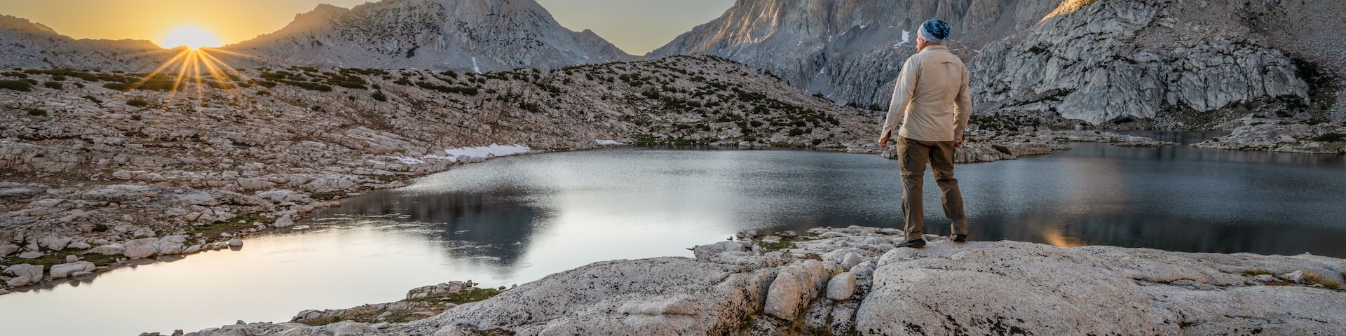 Rear view of a man standing on a rock in front of Grinnell lake at dawn looking at the Sierra Nevada Mountains, Sierra National Forest, California, USA