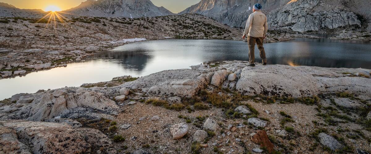 Rear view of a man standing on a rock in front of Grinnell lake at dawn looking at the Sierra Nevada Mountains, Sierra National Forest, California, USA