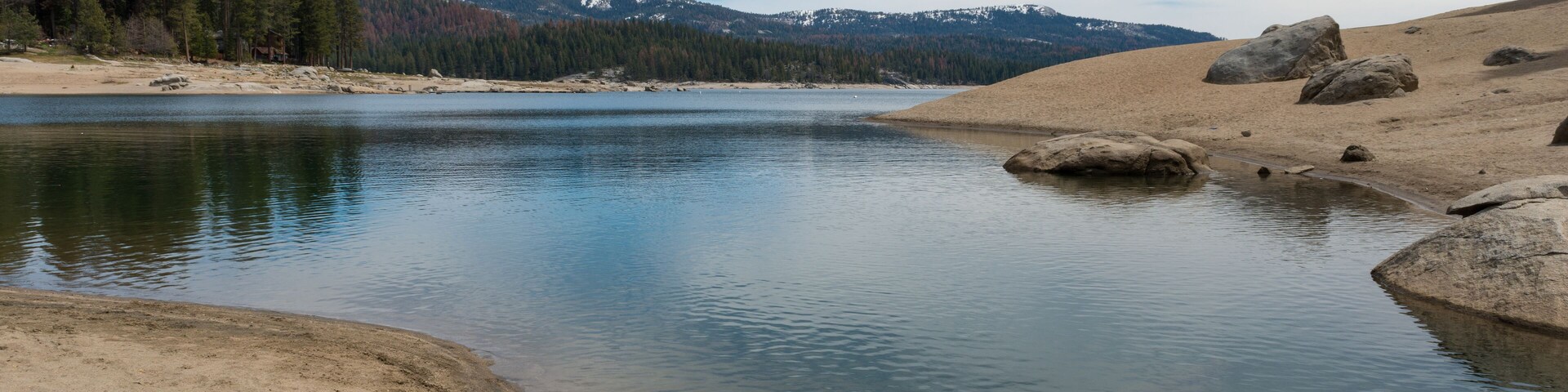 Um lago tranquilo rodeado por dunas de areia e uma cadeia montanhosa arborizada.