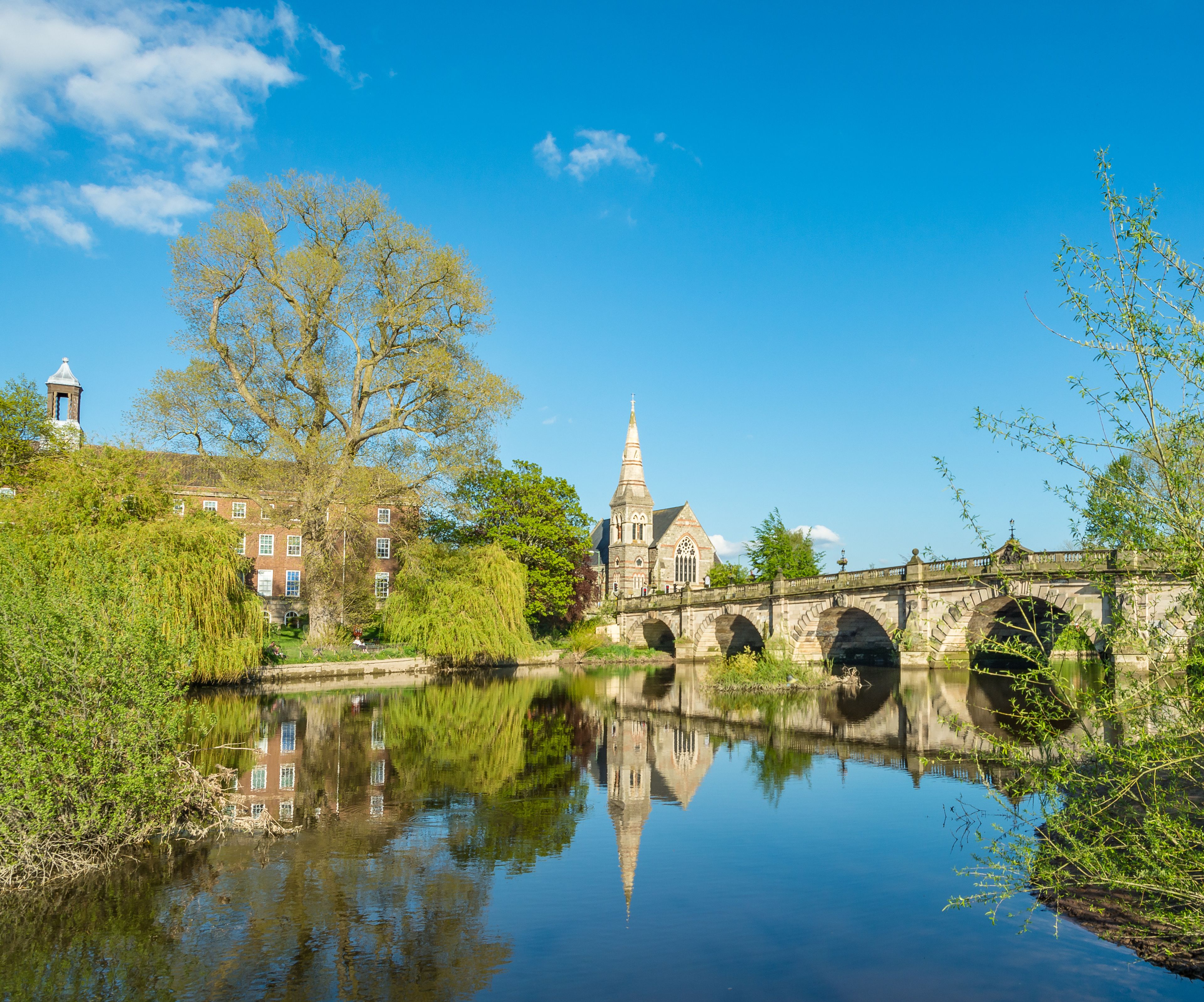 Une rivière paisible où se reflète un pont historique et une église coiffée d’une flèche.