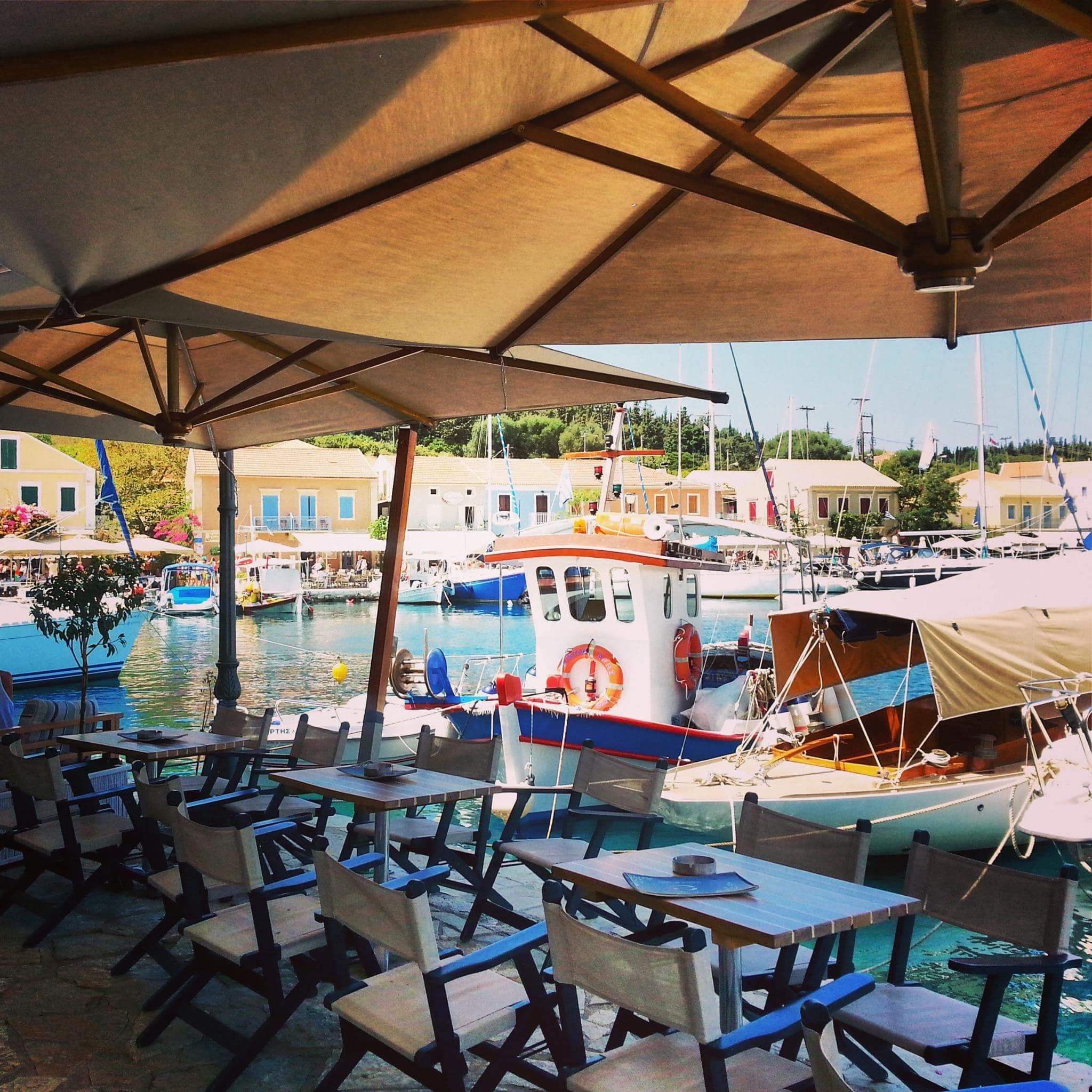 A marina with boats docked, tables and chairs under a canopy, and buildings in the background.