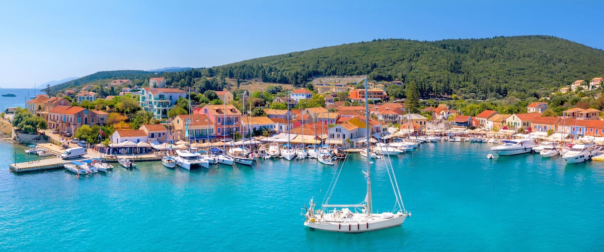 A coastal town with colorful buildings, a marina with boats, and a forested hill in the background.