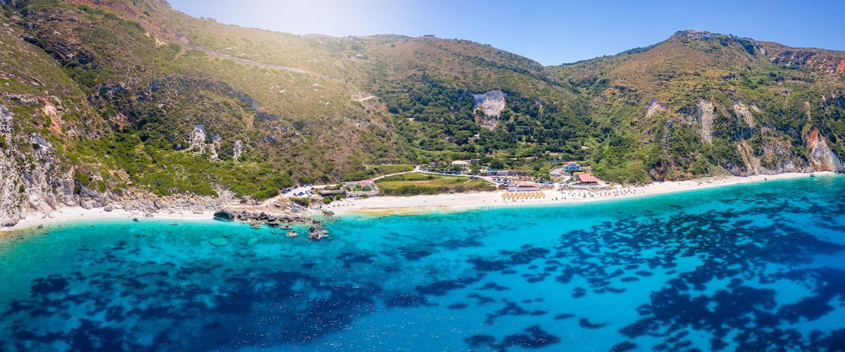 A coastal landscape with a beach, clear turquoise waters, and a mountainous backdrop.
