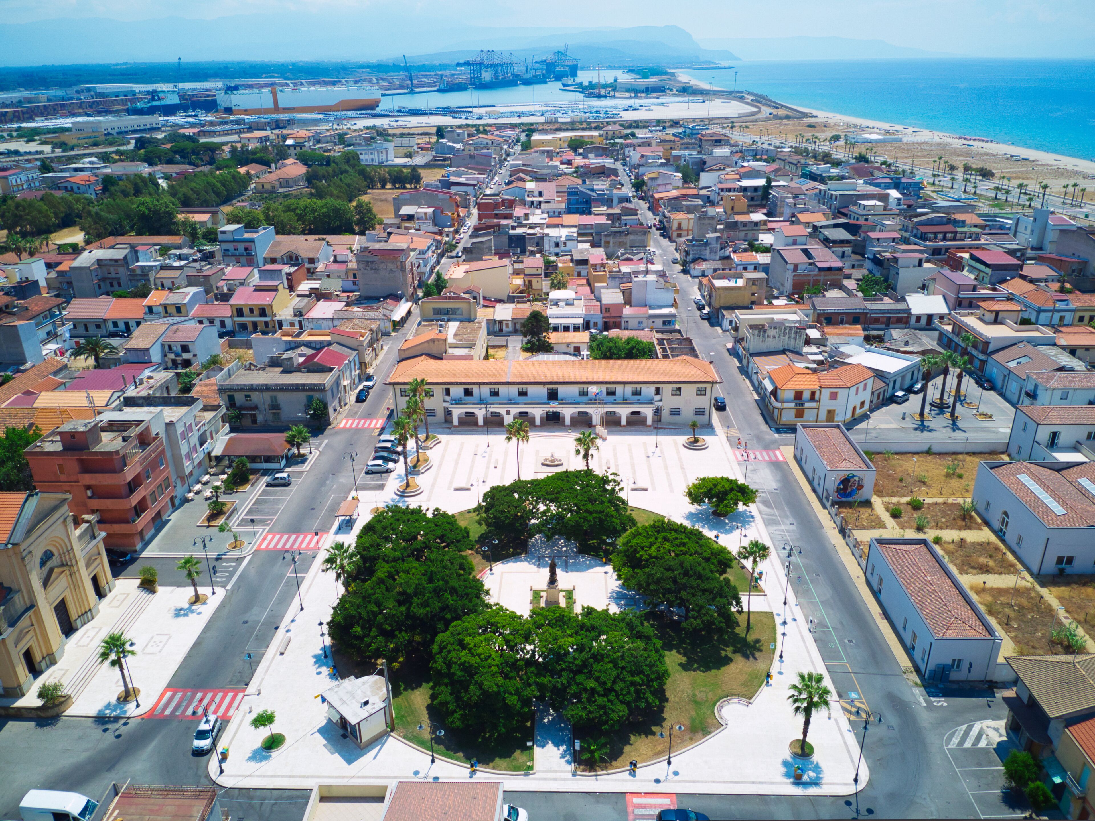 An aerial view of a coastal city with a central plaza surrounded by buildings, a beach, and a port with cranes.