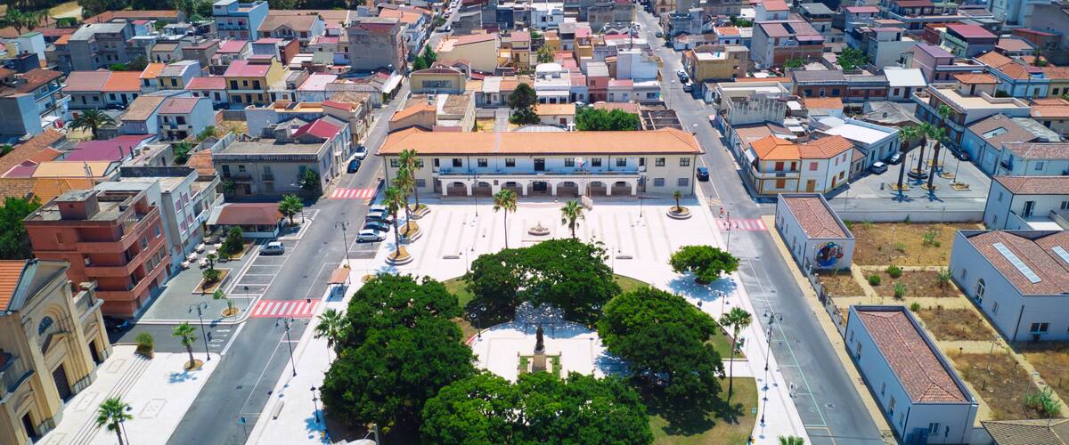 An aerial view of a coastal city with a central plaza surrounded by buildings, a beach, and a port with cranes.