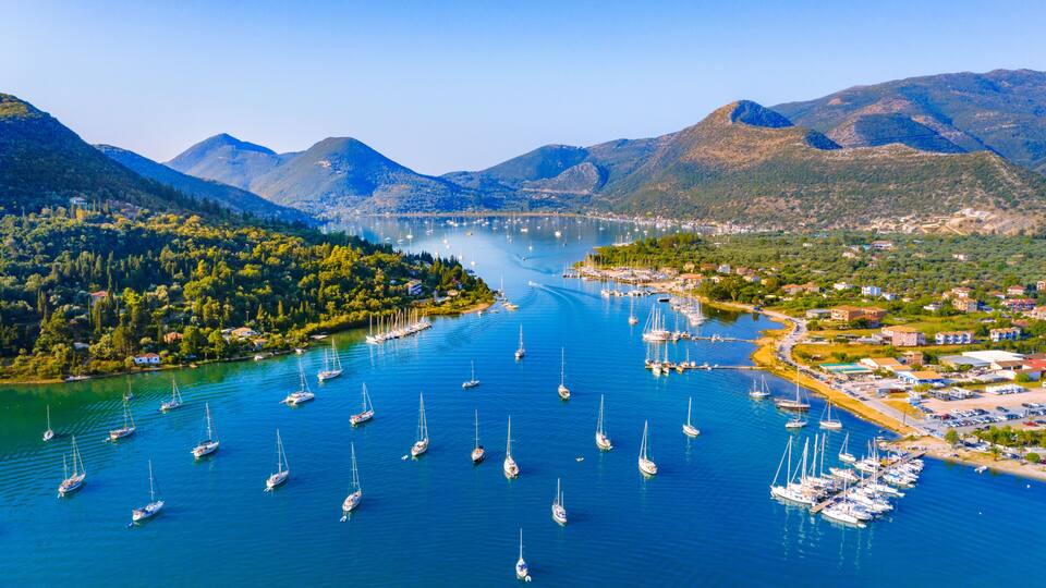 A lake with numerous sailboats, surrounded by mountains and a small town with buildings and greenery.