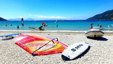 A pebbled beach with windsurfing equipment and a sailboat in the water.
