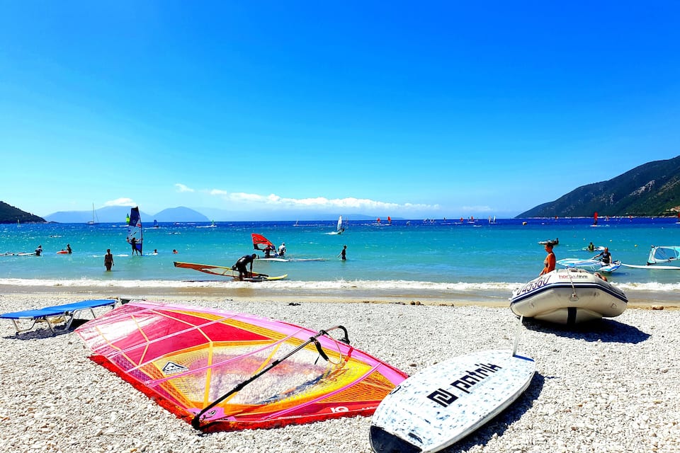 A pebbled beach with windsurfing equipment and a sailboat in the water.