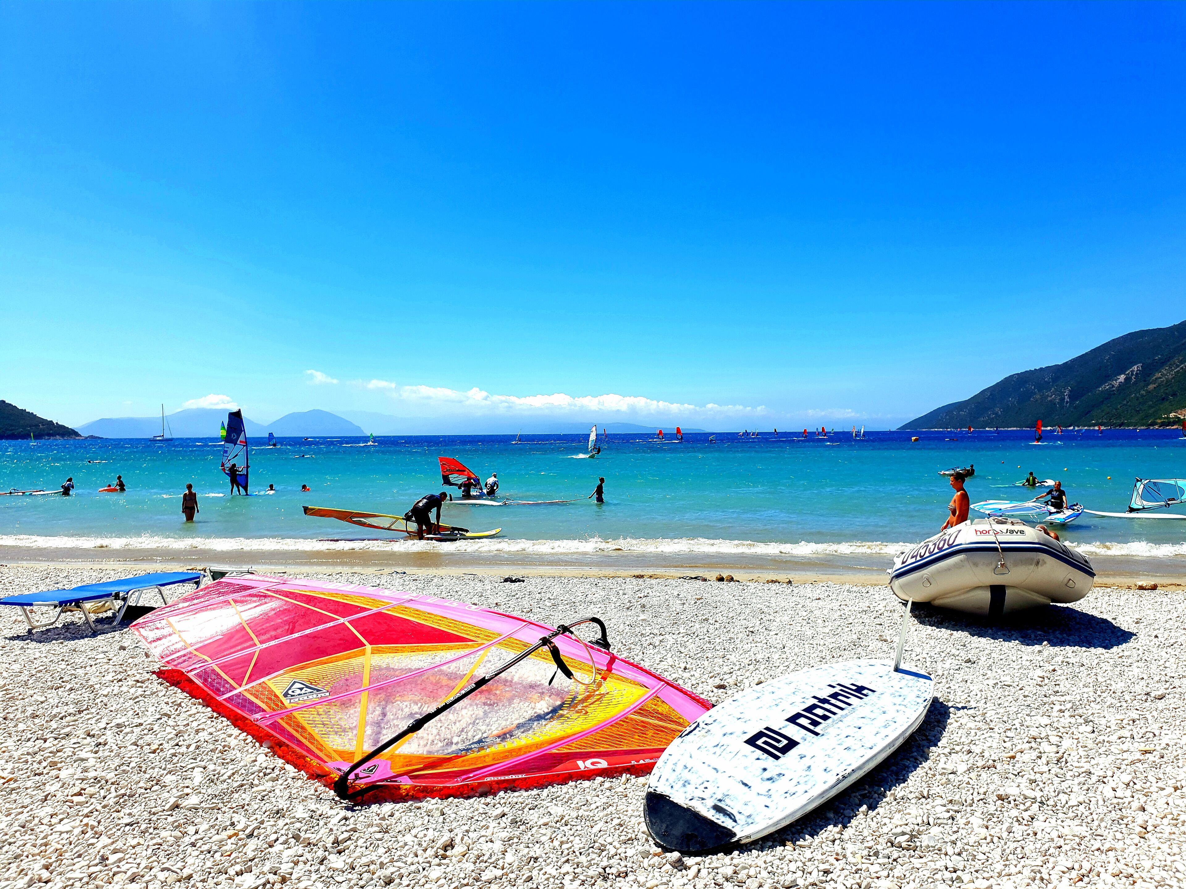 A pebbled beach with windsurfing equipment and a sailboat in the water.