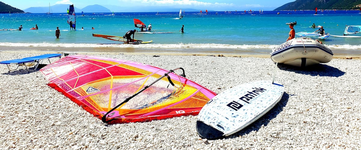 A pebbled beach with windsurfing equipment and a sailboat in the water.