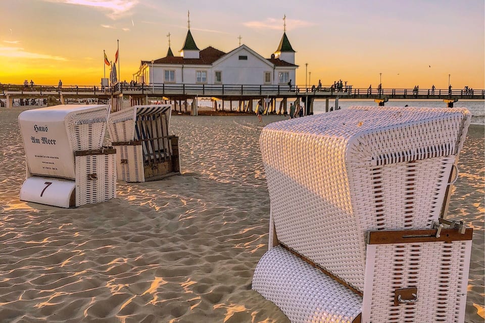 Une plage avec des chaises en osier blanches et une jetée sur laquelle se trouve un bâtiment blanc, avec des toits verts en arrière-plan.