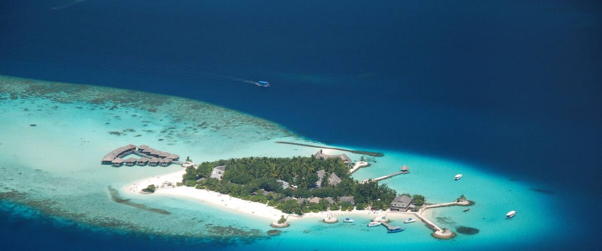 An aerial view of a tropical island with a white sandy beach, lush greenery, and several small buildings.