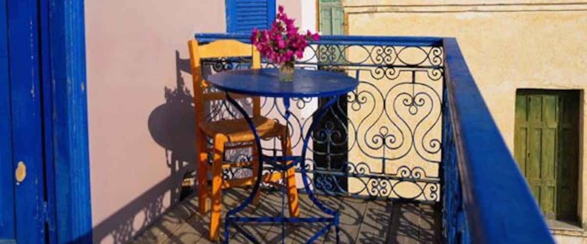 A blue door, a blue shutter, a table with chairs, a vase with pink flowers, and a building with a clock tower in the background.