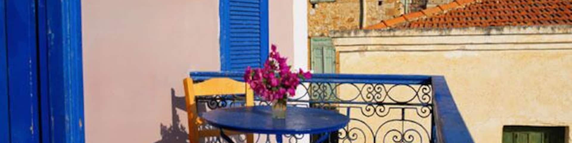 A blue door, a blue shutter, a table with chairs, a vase with pink flowers, and a building with a clock tower in the background.