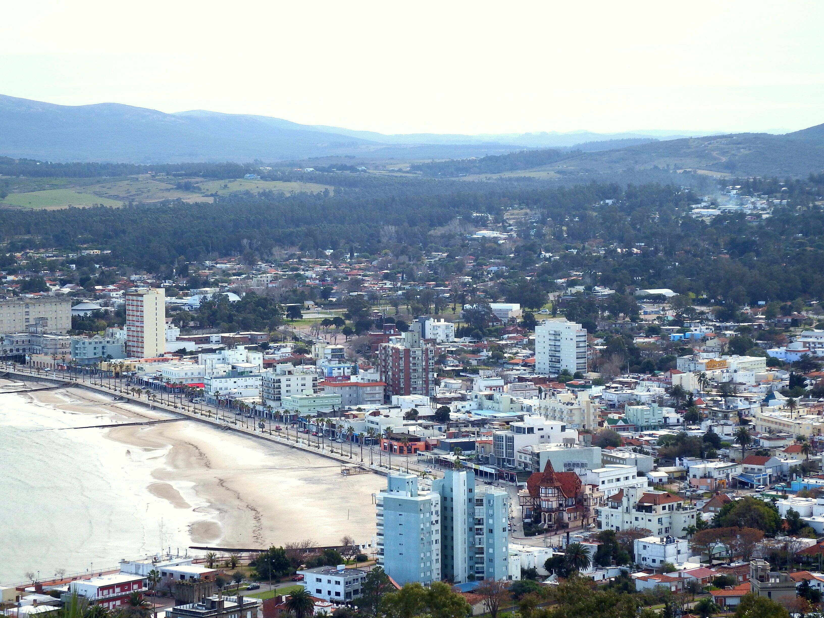 A coastal cityscape with a beach, buildings, and mountains in the background.