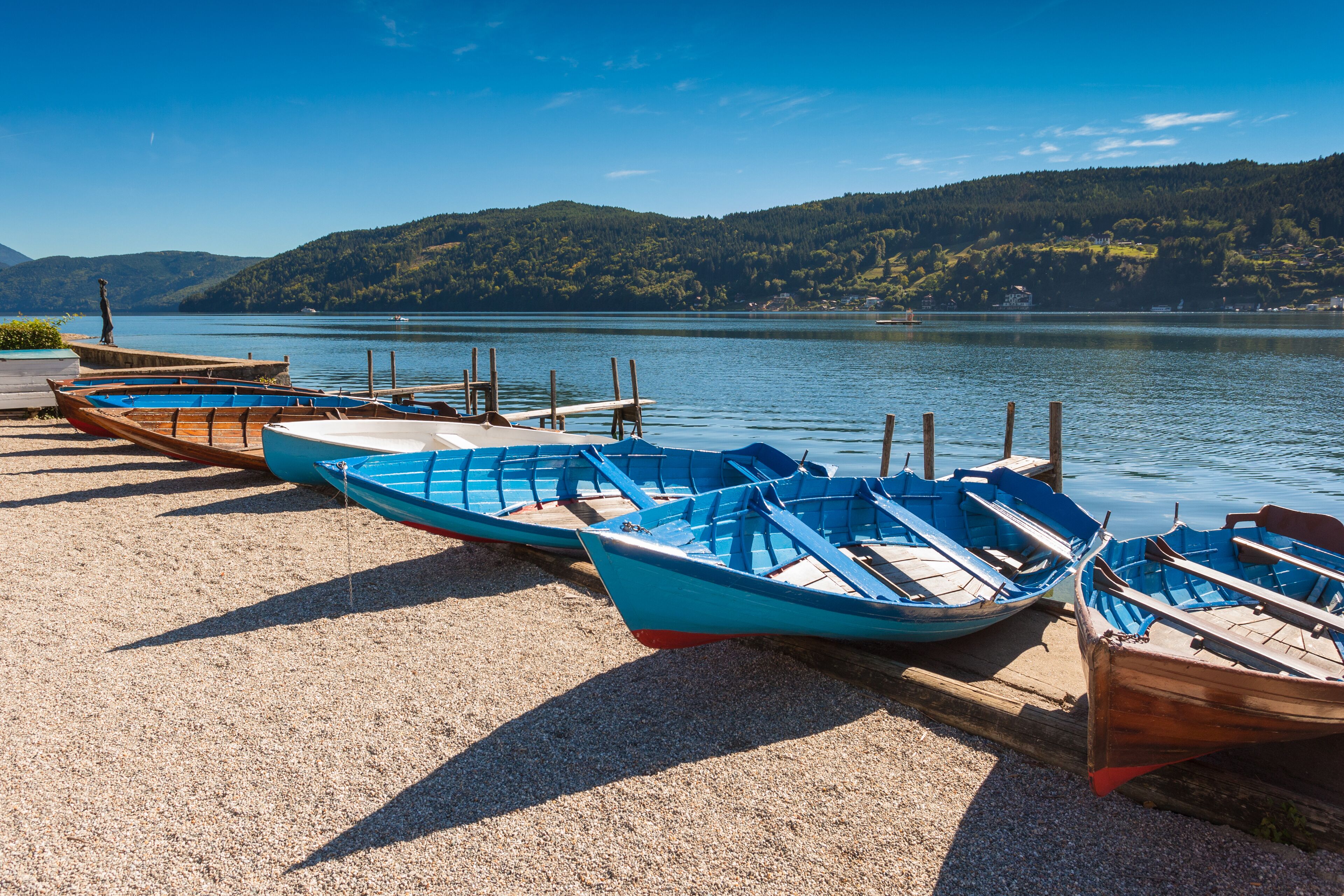 A row of wooden boats on a gravel beach by a calm lake with hills in the background.