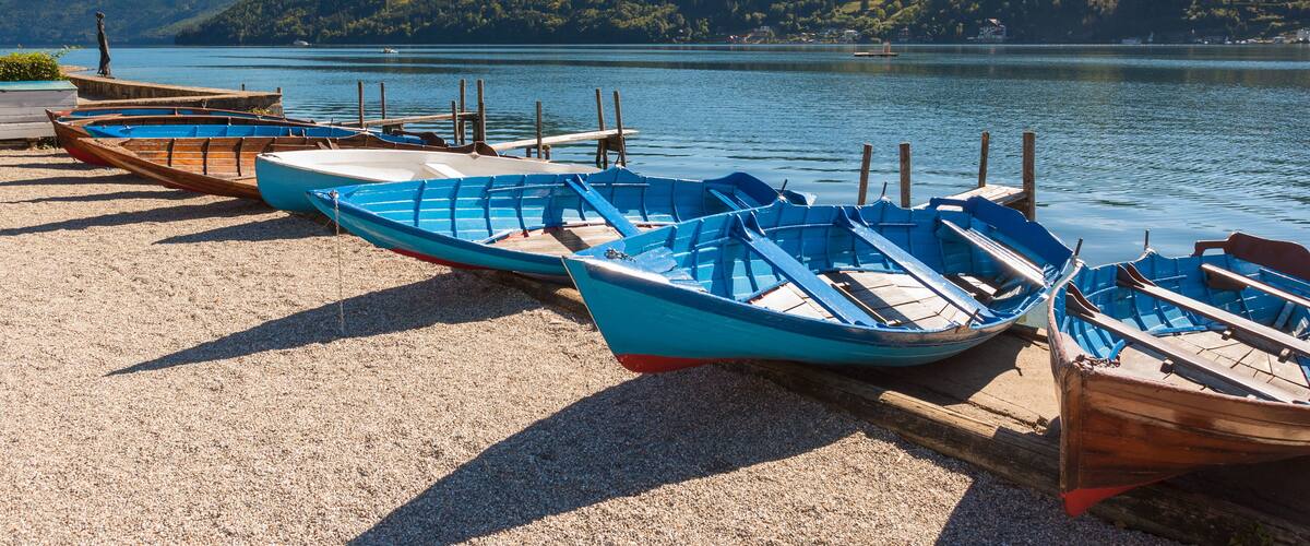 A row of wooden boats on a gravel beach by a calm lake with hills in the background.