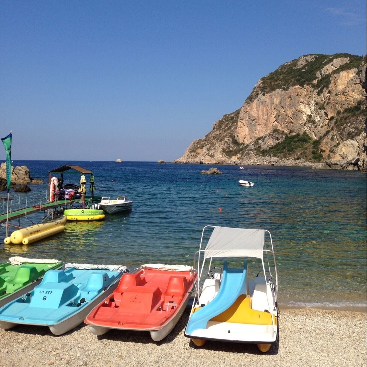 A beach with several small boats lined up on the shore, a rocky cliff in the background, and calm blue water.