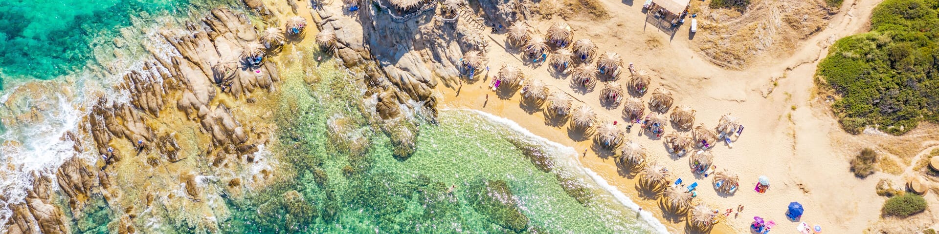 A coastal landscape with clear turquoise waters, rocky cliffs, and a sandy beach with thatched-roof structures.