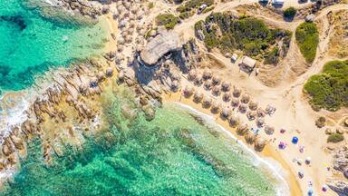 Paisagem costeira com águas turquesa cristalinas, falésias rochosas e uma praia de areia com estruturas de telhado de palha.