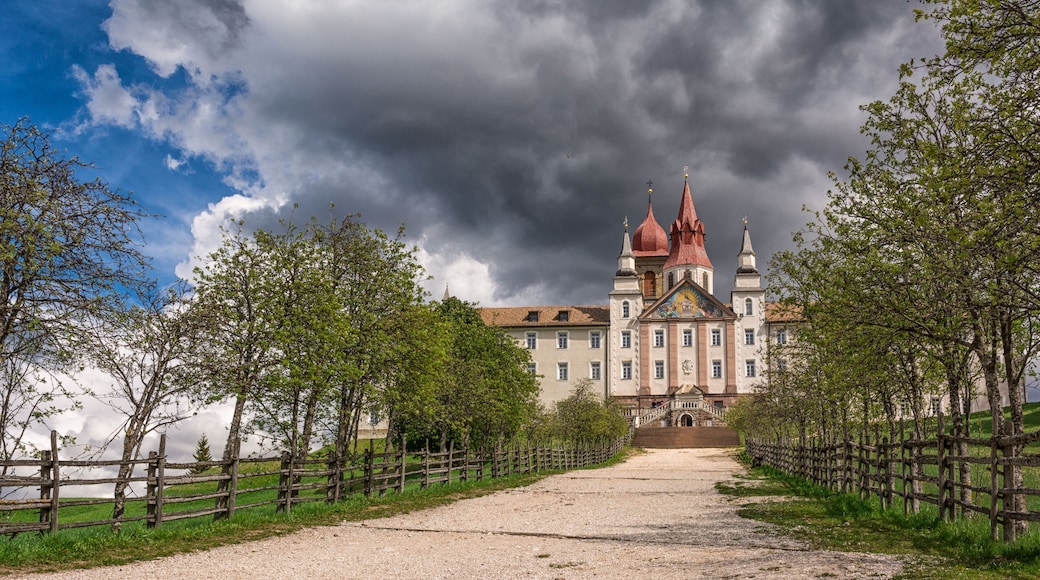 A historic building with multiple red spires, surrounded by a gravel path and wooden fences, under a cloudy sky.