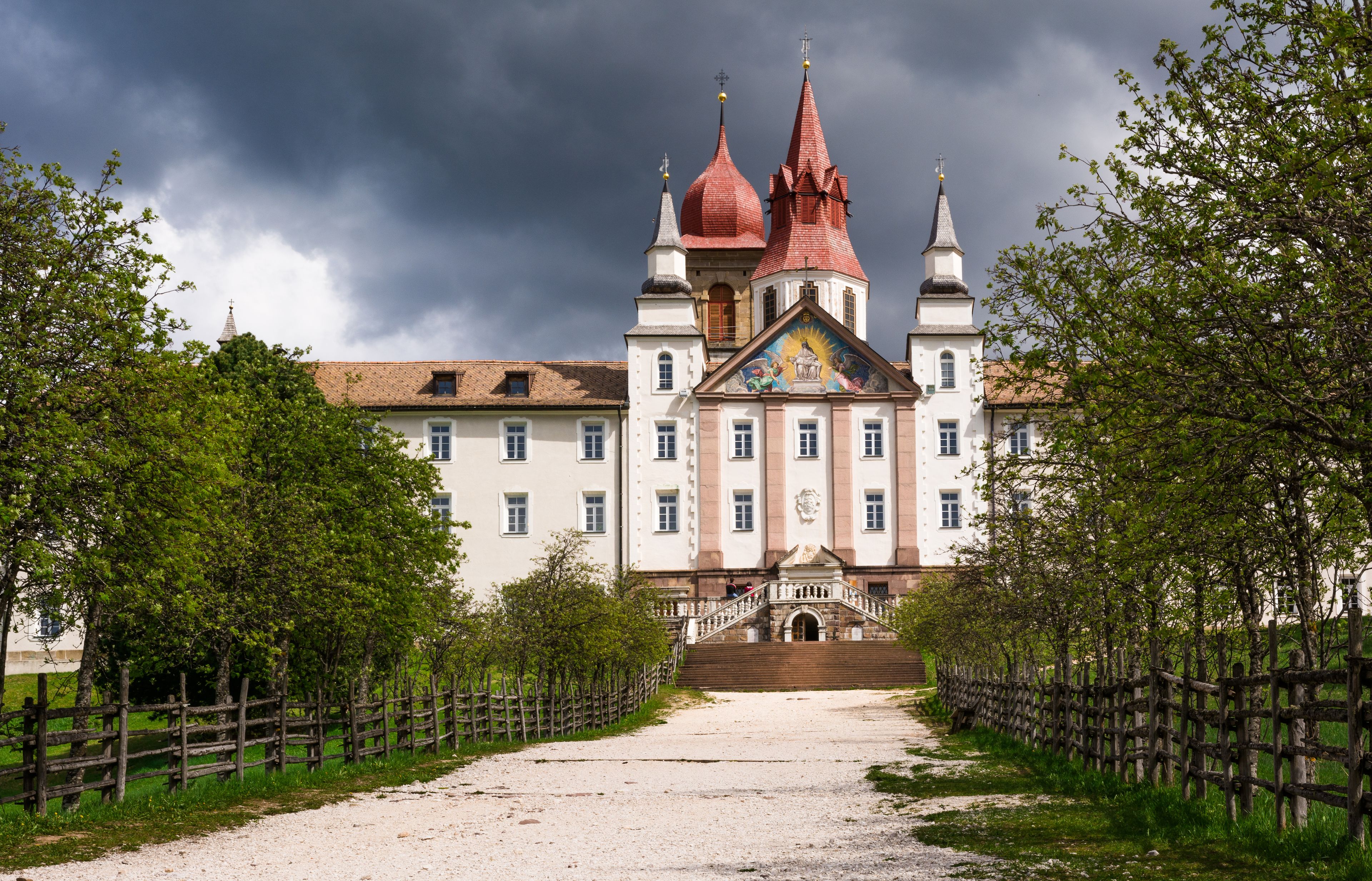 A historic building with a red-tiled roof and multiple spires, surrounded by a wooden fence and trees.