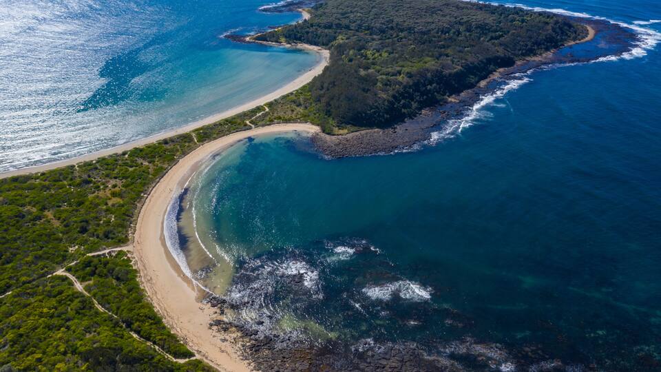 An aerial view of a coastal landscape with a sandy beach, a small island, and clear blue waters.