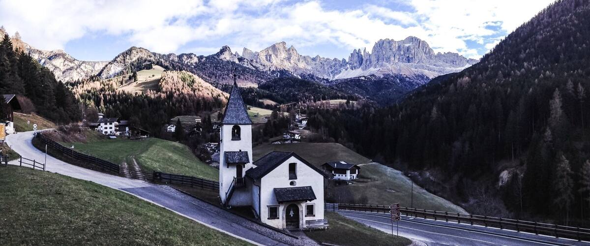 A church with a bell tower, surrounded by a winding road, mountains, and a forested valley.