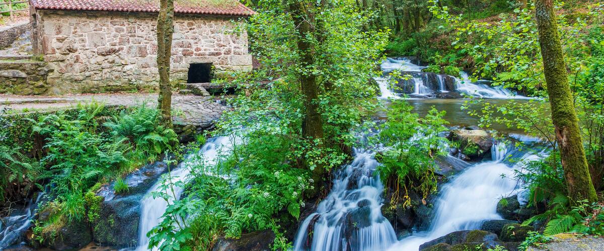 A stone building with a red roof, a waterfall, and lush greenery.
