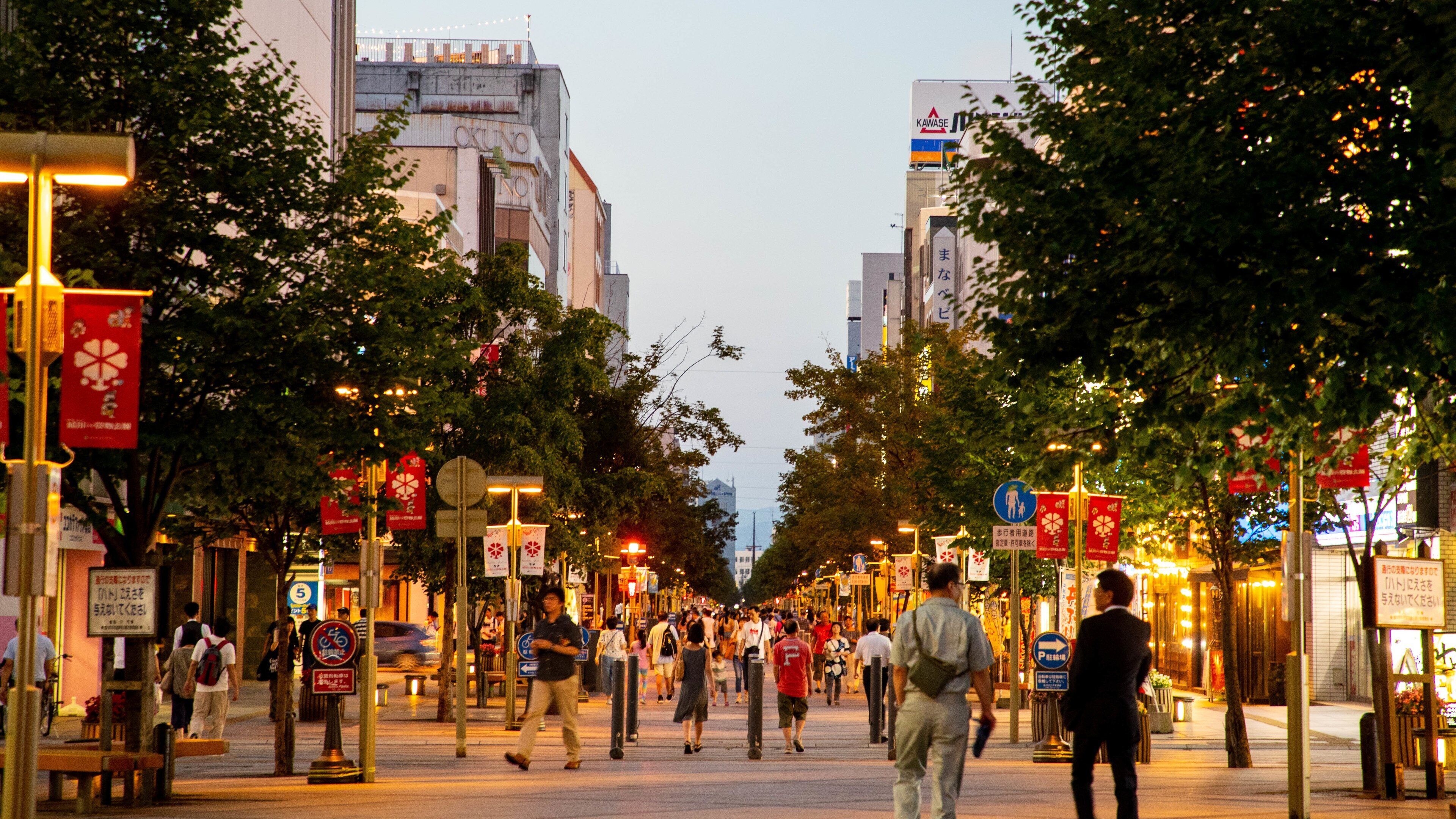 A busy urban street lined with trees and various shops.