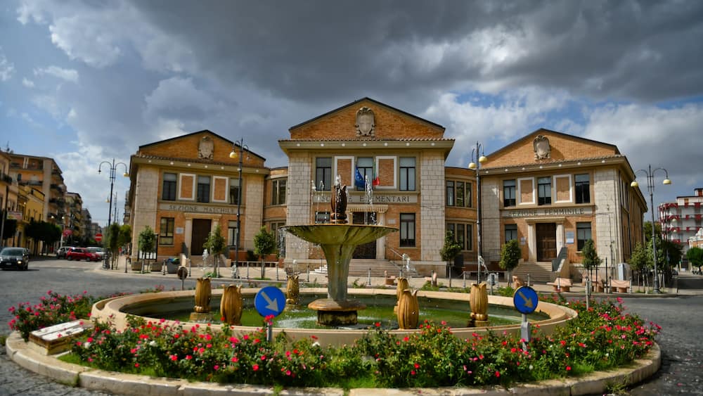 Une place publique avec une fontaine, entourée de parterres de fleurs et de deux bâtiments portant les inscriptions « SECTION MASCULINE » et « SECTION FÉMININE ».