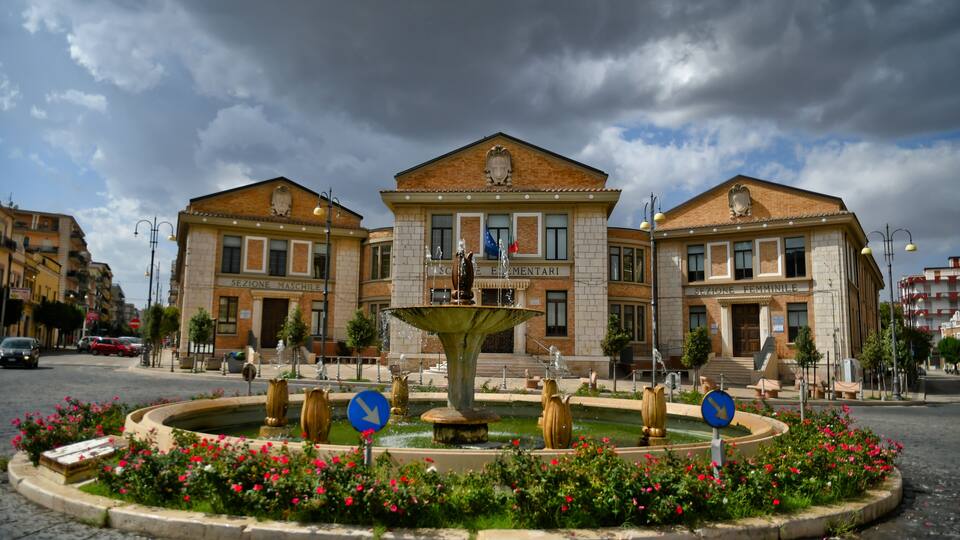 A public square with a fountain, surrounded by flower beds and two buildings labeled "SEZIONE MASCHILE" and "SEZIONE FEMMINILE".