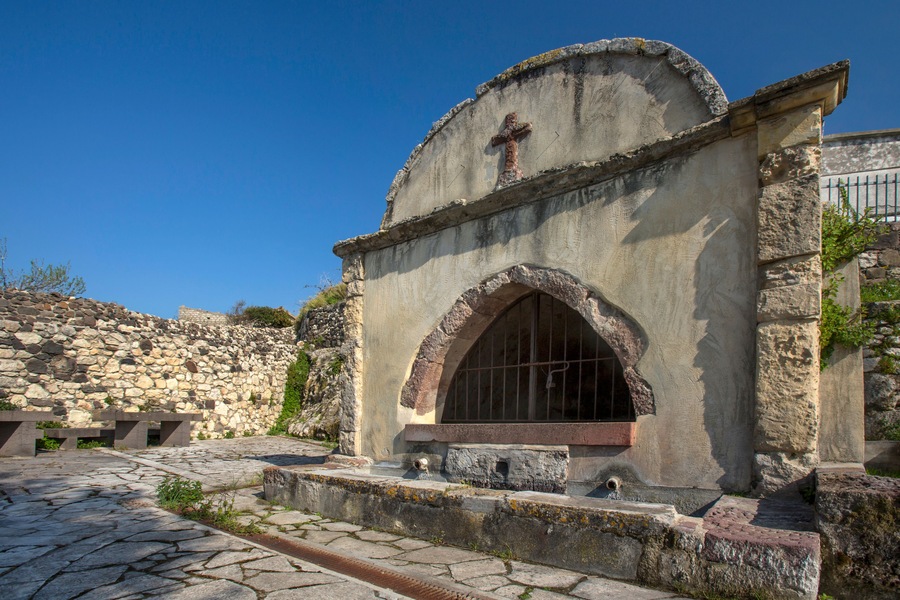 Una iglesia de piedra con una cruz, una entrada arqueada y un muro de piedra al fondo.