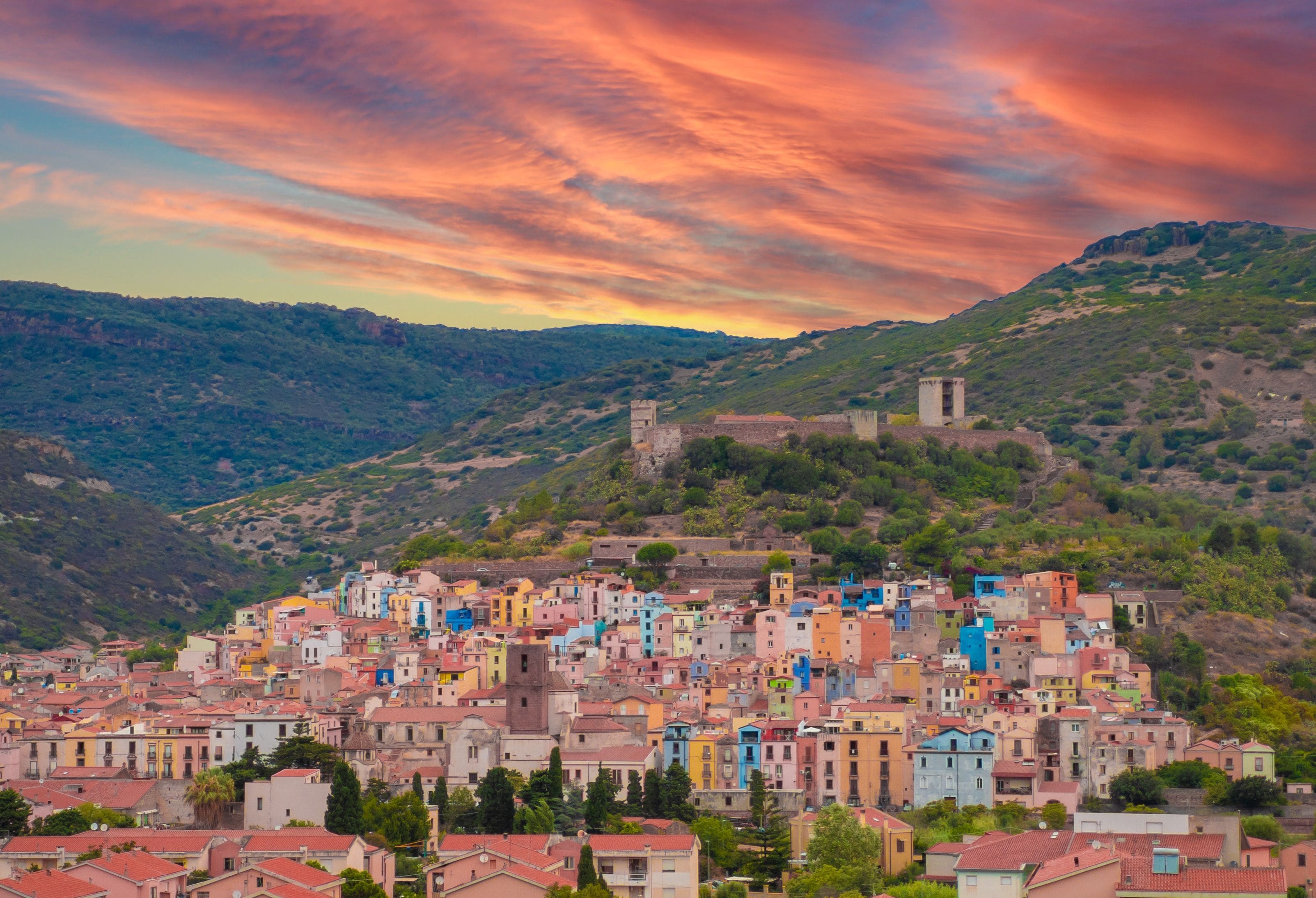 Una cittadina costiera con edifici colorati, un'imponente struttura in cima a una collina e un cielo al tramonto suggestivo.