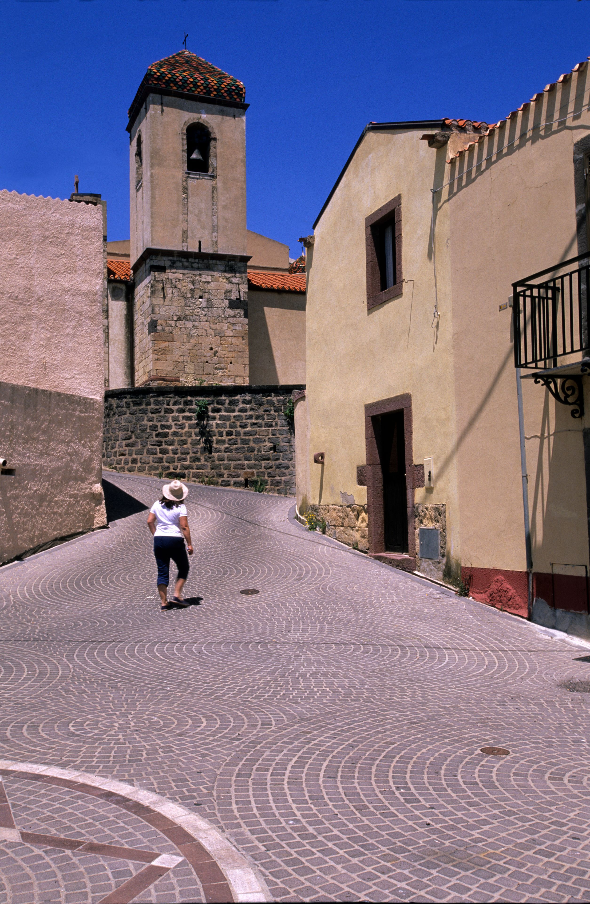 Una strada acciottolata con una donna che cammina, una torre campanaria e edifici dall'architettura tradizionale.